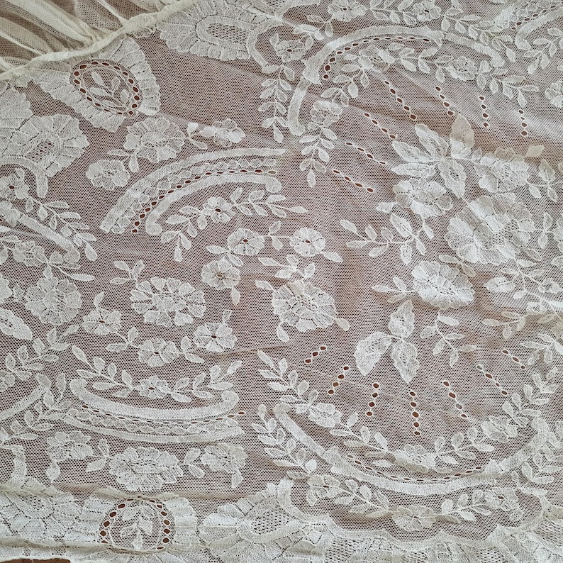 May include: Detailed view of white lace fabric with floral and leaf motifs. The lace showcases elaborate embroidery and openwork, giving a textured and airy feel. The fabric is likely used for decorative purposes, such as clothing or home decor.