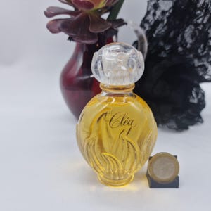 Puede incluir: Un frasco de perfume de vidrio redondo lleno de líquido dorado, con la palabra Clea en letra cursiva en el frente. El frasco tiene un tapón transparente facetado y una base texturizada. Una moneda está colocada al lado para dar escala.
