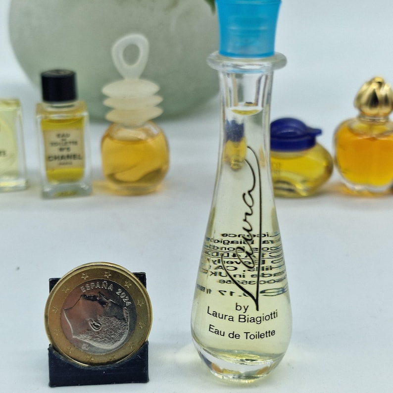 K&ouml;nnte beinhalten: Eine Sammlung von Miniatur-Parf&uuml;mflaschen, darunter eine klare Flasche mit blauem Verschluss, die mit "Laura Biagiotti Eau de Toilette" beschriftet ist. Andere Flaschen haben gelbe und schwarze Verschl&uuml;sse. Eine M&uuml;nze aus Spanien ist im Vordergrund.