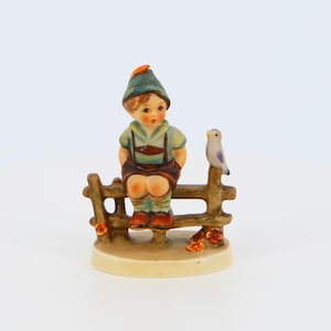 Puede incluir: Una figurita de porcelana de un niño con un sombrero verde y naranja sentado en una valla de madera con un pequeño pájaro azul posado en un poste de la valla.
