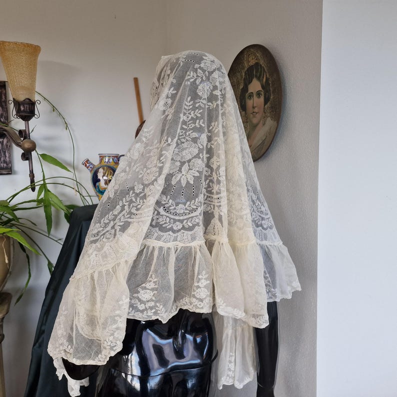 May include: An antique ivory lace mantilla, with detailed floral and leaf patterns. The delicate fabric is draped over a mannequin, highlighting the intricate embroidery and ruffled edges. A traditional head covering.