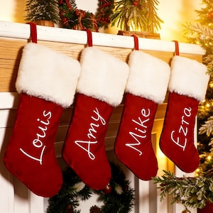 Könnte beinhalten: Vier rote Weihnachtsstrümpfe mit weißen Kunstfellbündchen hängen an einem Kaminsims. Jeder Strumpf ist mit einem Namen in weißer Schrift personalisiert: Louis, Amy, Mike und Ezra. Festliche Weihnachtsdekoration umgibt die Strümpfe.