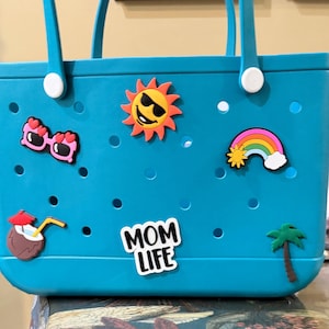 May include: A teal rubber tote bag featuring a perforated design and two handles. The bag is decorated with colorful charms including heart-shaped sunglasses, a smiling sun, a rainbow, a coconut drink, a palm tree, and text reading MOM LIFE.