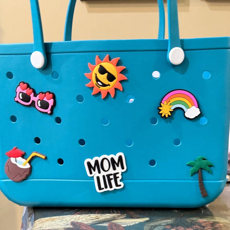 May include: A teal rubber tote bag featuring a perforated design and two handles. The bag is decorated with colorful charms including heart-shaped sunglasses, a smiling sun, a rainbow, a coconut drink, a palm tree, and text reading MOM LIFE.