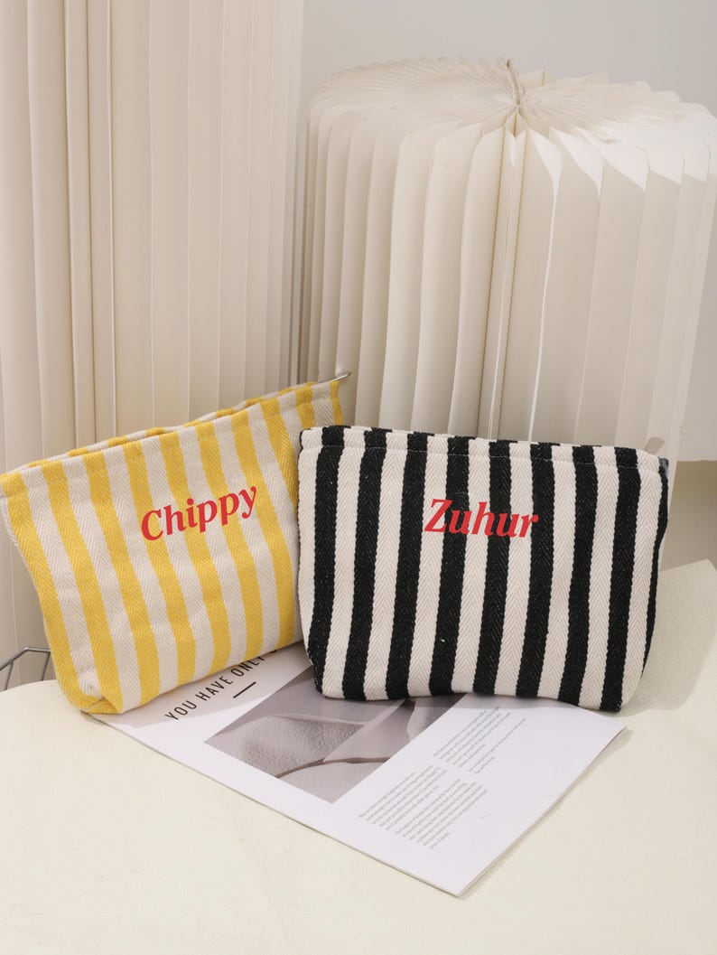 May include: Two striped fabric pouches with zipper closures. The pouch on the left is yellow and white with the word "Chippy" embroidered in red. The pouch on the right is black and white with the word "Zuhur" embroidered in red.
