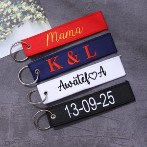 May include: Four embroidered keyrings in red, navy blue, white, and black. The red keyring says "Mama" in yellow script. The navy blue keyring has "K & L" in orange. The white keyring says "Awatef ♡ A". The black keyring says "13-09-25".