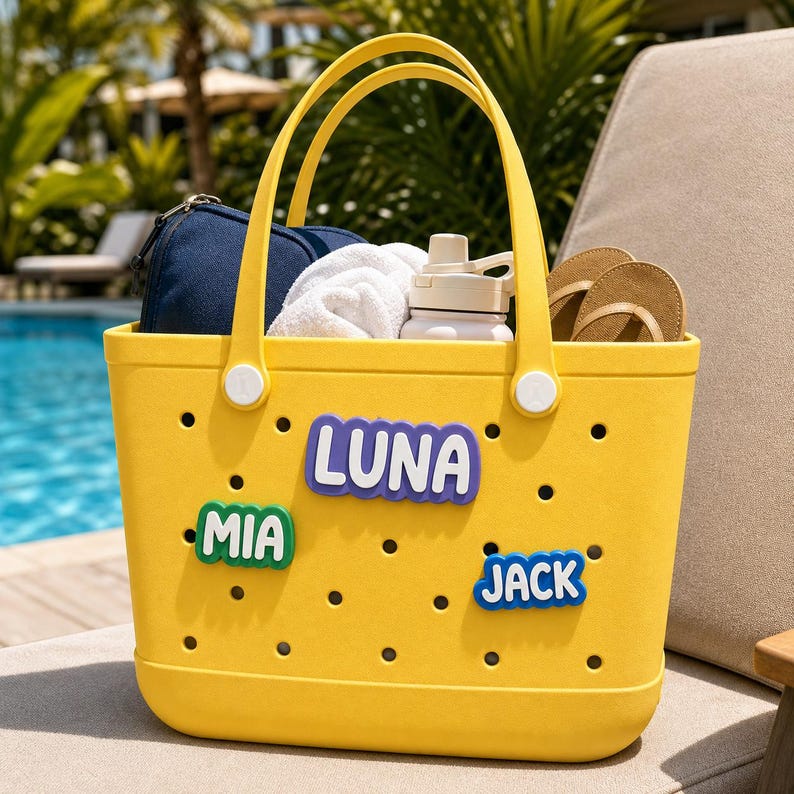 May include: A bright yellow perforated tote bag featuring three colorful name charms attached to the front. The charms display the names LUNA in purple, MIA in green, and JACK in blue. The bag is filled with a dark blue pouch, a white towel, a white water bottle, and tan flip-flops.