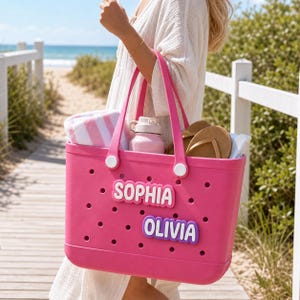 May include: A bright pink rubber tote bag featuring a perforated design and two white circular accents on the handles. The bag is customized with two rectangular name tags in white and purple, displaying the names SOPHIA and OLIVIA in bold letters.