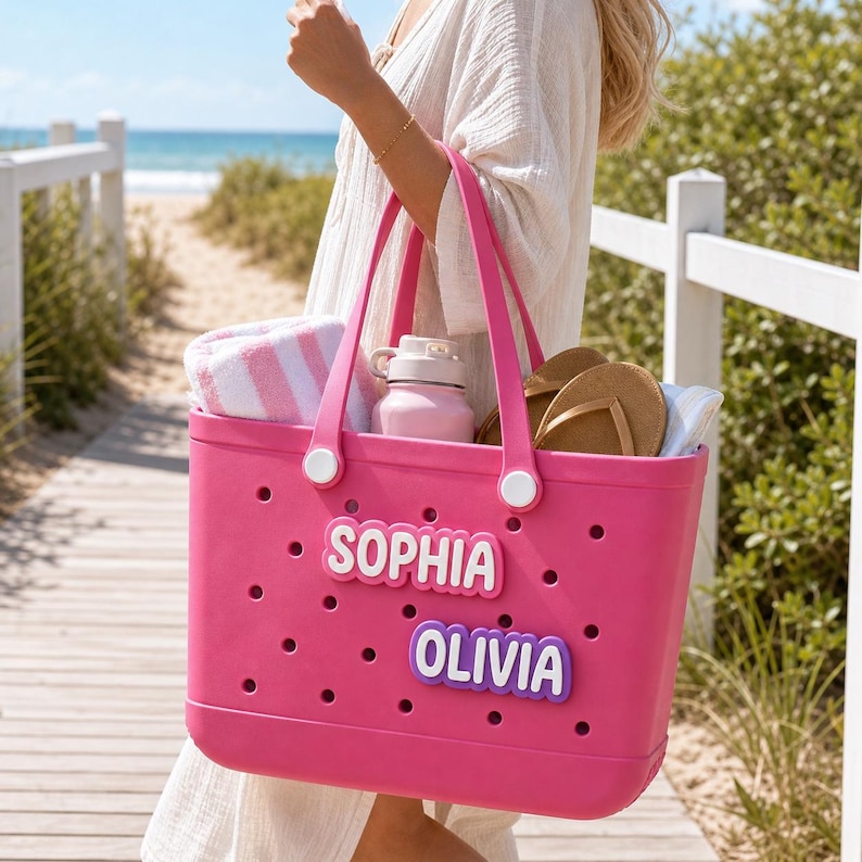 May include: A bright pink rubber tote bag featuring a perforated design and two white circular accents on the handles. The bag is customized with two rectangular name tags in white and purple, displaying the names SOPHIA and OLIVIA in bold letters.