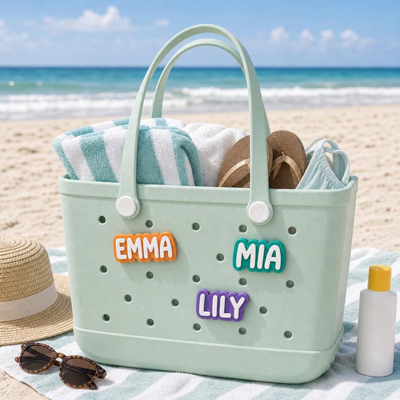 May include: A light green perforated rubber tote bag featuring three colorful name charms that read EMMA in orange, MIA in teal, and LILY in purple. The bag is filled with a white and teal striped towel, brown flip-flops, and a light blue garment.
