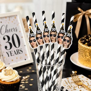 Op de afbeelding: Een verzameling zwart-wit gestreepte papieren rietjes met feesthoedjes met een vrouw met een zonnebril en het nummer 30. De rietjes staan in een glas, met een ingelijst bord met de tekst "Cheers 30 YEARS".