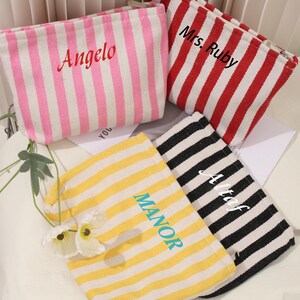 May include: Four striped canvas zippered pouches with different names embroidered on them. The pouches are pink and white, red and white, yellow and white, and black and white.