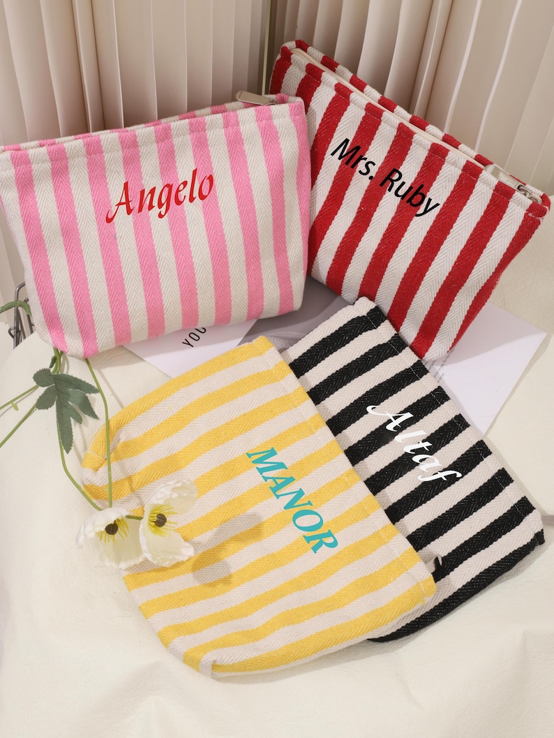 May include: Four striped canvas zippered pouches with different names embroidered on them. The pouches are pink and white, red and white, yellow and white, and black and white.