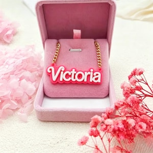 Puede incluir: Un collar rosa con el nombre "Victoria" en letras blancas, presentado en una caja de joyería rosa. El collar tiene una cadena dorada. Flores rosas en el fondo.