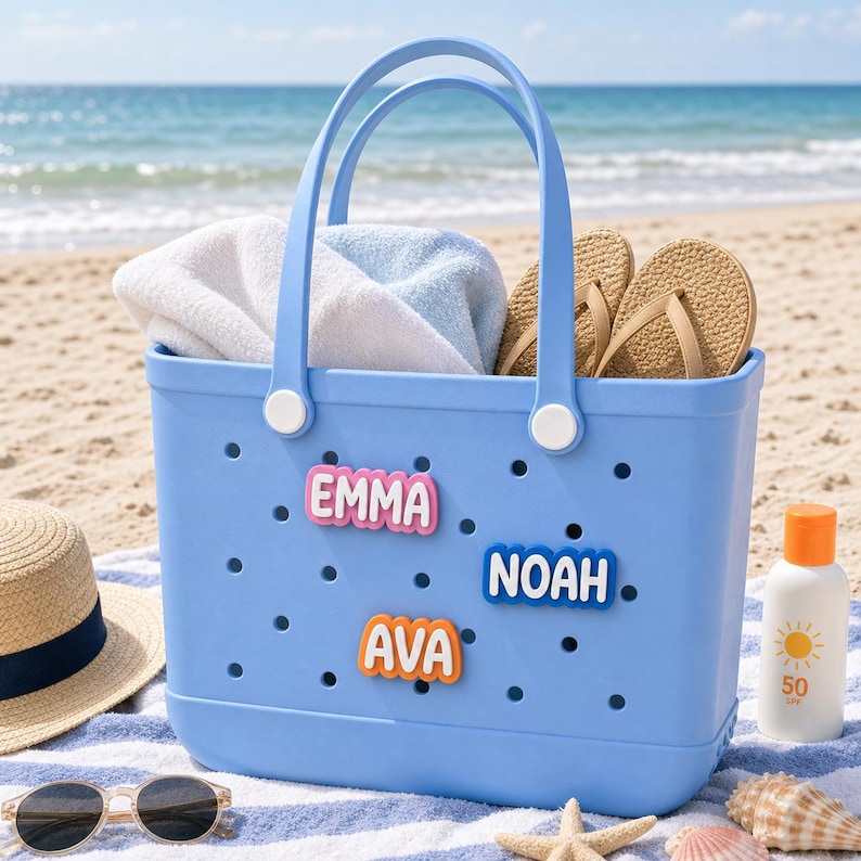 May include: A light blue perforated rubber tote bag featuring three colorful name charms attached to the front. The charms display the names EMMA in pink, NOAH in blue, and AVA in orange. The bag has two matching blue handles and white rivets.