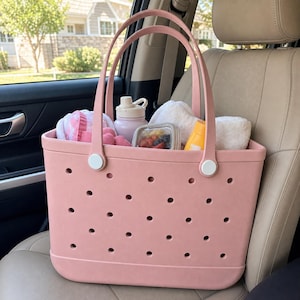 May include: A dusty pink tote bag featuring a perforated pattern across the body, two tall handles attached with white circular hardware, and a spacious interior filled with a light pink water bottle, a food container, and white towels.