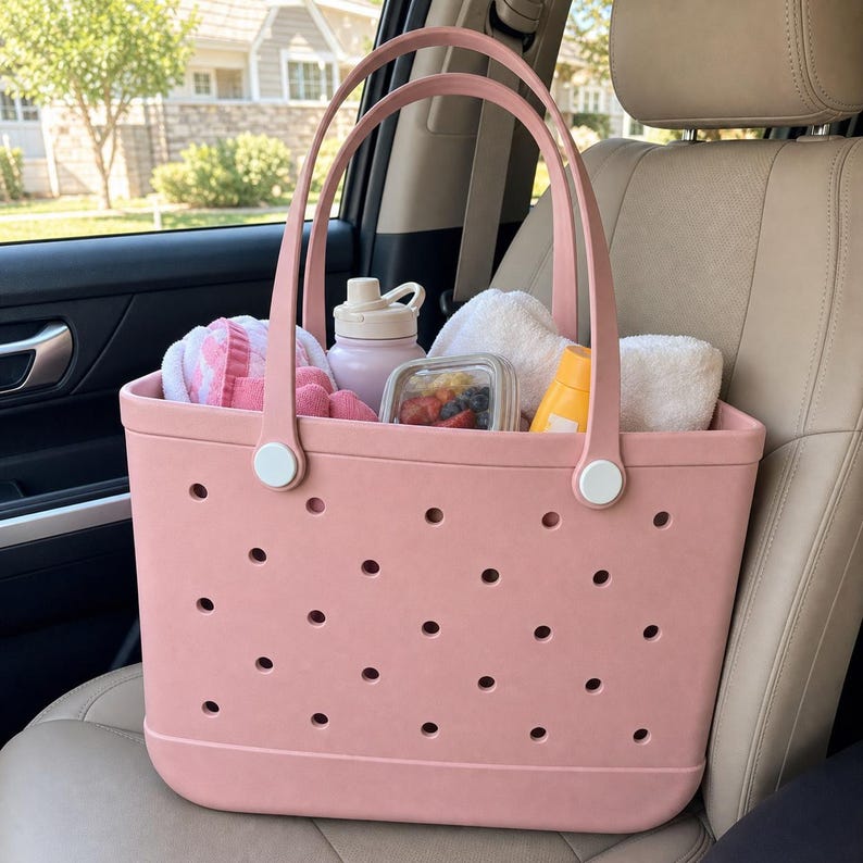 May include: A dusty pink tote bag featuring a perforated pattern across the body, two tall handles attached with white circular hardware, and a spacious interior filled with a light pink water bottle, a food container, and white towels.