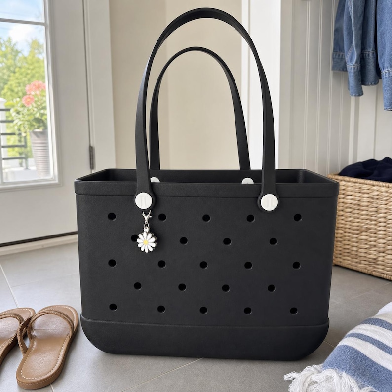 May include: A black rectangular tote bag featuring a perforated body with evenly spaced circular holes. The bag has two tall black handles attached with white circular hardware. A small white and yellow daisy charm hangs from one handle.