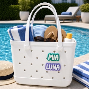 May include: A white perforated tote bag featuring two handles and two colorful rubber charms attached to the front. The top charm is teal with the white text MIA, and the bottom charm is purple with the white text LUNA. The bag contains towels, sandals, and sunglasses.