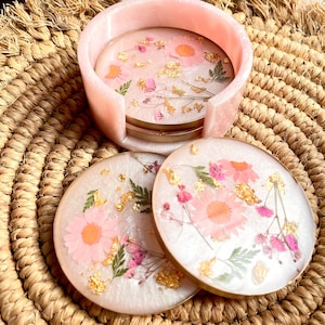 May include: Set of four pink and white coasters with pressed flowers and gold leaf accents. The coasters are arranged in a circle and are sitting on a woven straw mat. The coasters are in a pink holder.