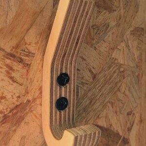 May include: A wooden coat hook with a curved design, made from layered plywood. The hook is attached to a wooden surface with two black screws. The wood has a natural, light brown colour.