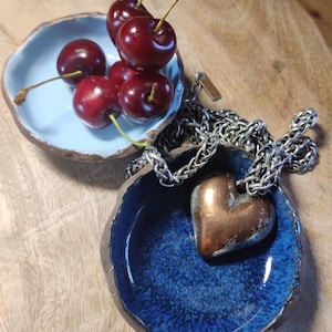 May include: A still life featuring two small, handmade ceramic bowls. One is light blue and holds cherries, the other is dark blue and contains a copper heart pendant on a silver chain. The bowls sit on a wooden surface.