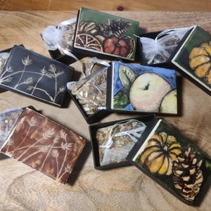 May include: A collection of small black boxes with hand-painted lids. The lids display images of pumpkins, apples, pine cones, and wheat. The boxes are open, revealing small bags of contents, presented on a wooden surface.