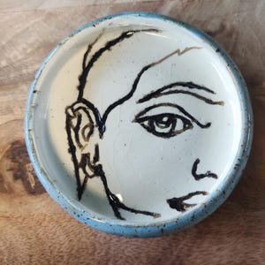 May include: A small, circular ceramic dish with a pale blue rim and a cream interior. The dish has a black line drawing of a human face, including an eye, ear, and part of the face. The dish is on a wooden surface.