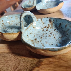 May include: Handmade ceramic serving dishes with a light blue speckled glaze and a natural, unglazed rim. One dish has a handle. The dishes are on a wooden surface. These unique bowls are perfect for serving snacks or dips.