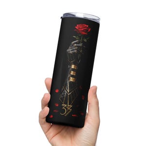 Puede incluir: Vaso de acero inoxidable negro con un brazo robótico estilizado sosteniendo una rosa roja. Acentos dorados y el número 33 son visibles en el vaso. Pétalos de rosa rojos esparcidos alrededor de la base.