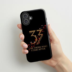 Puede incluir: Funda de teléfono negra con el número "33" en dorado y detalles de corazones rojos. El texto "FOR THOSE WHO COME AFTER" está debajo. La funda está sujeta en una mano.