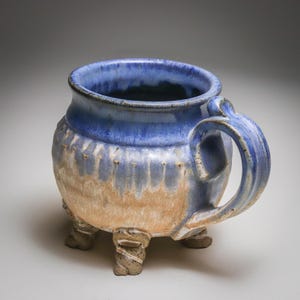 May include: A ceramic mug with a rounded body and three short, textured feet. The upper portion features a deep blue glaze that drips down into a light tan, earthy base. It includes a curved handle and a wide, circular opening at the top.