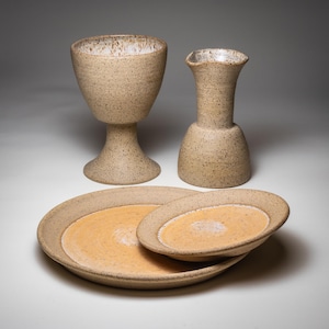 May include: A set of speckled ceramic tableware, including a chalice, a vase, and two plates. The plates have a light orange center and a speckled tan rim. The chalice and vase are tan with a speckled finish.