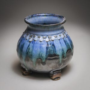May include: A round ceramic pot with three small feet, featuring a glaze that transitions from a light blue rim to a deep blue and turquoise drip pattern, ending in a dark grey base with a textured, stamped band around the upper neck area.