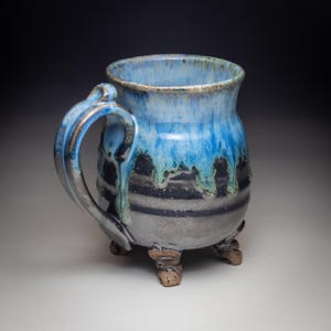 May include: A handmade ceramic mug with a blue and black glaze. The mug has a unique shape, a large handle, and sits on three small feet. The blue glaze drips down the sides, creating a textured effect. The mug is suitable for hot or cold beverages.
