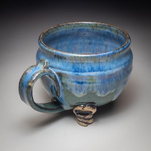 May include: A handmade ceramic mug with a blue glaze. The mug has a rounded shape, a handle, and three small feet. The blue glaze has variations in color, with lighter and darker shades. The mug is suitable for coffee, tea, or other beverages.