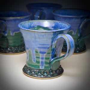 May include: Four handmade ceramic mugs with a blue and green glaze. The mugs have a unique design with a textured base and handle. The glaze drips down the sides, creating a visually appealing effect. These mugs are perfect for enjoying your favorite hot beverages.