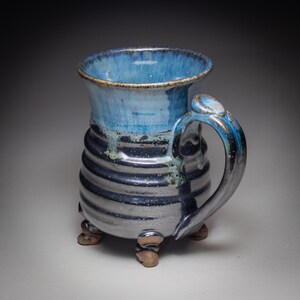 May include: A handmade ceramic mug with a blue and gray glaze. The mug has a cylindrical body with horizontal ridges, a rounded handle, and three small feet. The interior and rim are a lighter shade of blue.