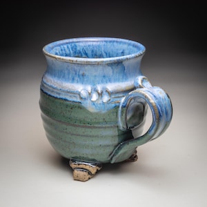 May include: Handmade ceramic mug with a blue and green glaze. The mug has a rounded body, a curved handle, and three small feet. The rim and handle are a lighter blue, while the body is a darker green. The mug is approximately 4 inches tall.