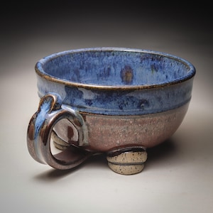 May include: A handmade ceramic bowl with a blue glazed interior and a reddish-brown exterior. The bowl has a handle and three small feet. The glaze has a mottled appearance, adding to the rustic charm of the piece.
