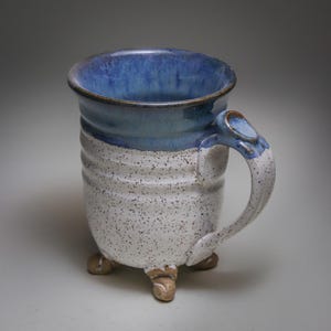May include: A ceramic mug featuring a speckled white body with horizontal ridges and a smooth, deep blue glazed rim. The mug stands on three small, light brown feet and includes a curved handle with a blue accent at the top attachment point.