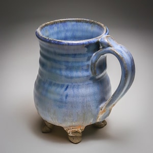 May include: A ceramic mug featuring a rounded body with a blue glaze that transitions to lighter tones. The mug has a sturdy handle on the side and stands on three small, textured feet. The rim is slightly flared, and the surface shows subtle ridges.