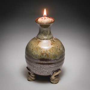 May include: A ceramic oil lamp with a rounded body and a narrow neck, featuring a glossy glaze that transitions from a speckled tan at the top to a dark brown at the base. The vessel stands on three small, textured, light-colored feet and holds a lit wick.