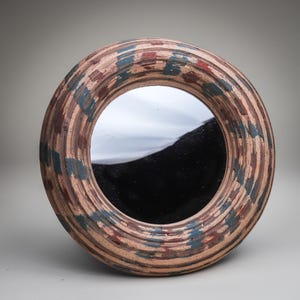 May include: A round wall mirror featuring a thick, textured frame with a light tan base color. The frame is decorated with repeating, short, horizontal brushstrokes in muted shades of dark blue, deep red, and brown, creating a layered pattern.