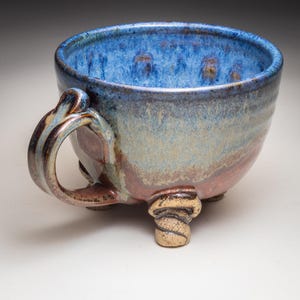 May include: A handmade ceramic mug with a blue interior and a textured exterior in shades of blue, brown, and tan. The mug has a large handle and three decorative feet. This unique mug is perfect for coffee or tea.