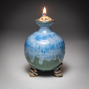 May include: A handmade ceramic oil lamp with a blue and green glaze. The lamp has a round body, a narrow neck, and three small feet. A lit wick sits atop the lamp, producing a warm glow. The lamp is a decorative and functional item.