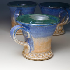 May include: Handmade ceramic mugs with a blue and tan glaze. The mugs have a unique shape with a wide rim and a decorative base. The handles are curved and comfortable to hold. The interior of the mugs is a deep green color.