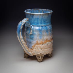 May include: A handmade ceramic mug with a blue and tan glaze. The mug has a large handle and sits on three small feet. The blue glaze is concentrated at the top and fades down the sides, with a tan band in the middle.