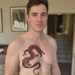 Puede incluir: Tatuaje de dragón rojo y negro en el pecho de una persona. El dragón está estilizado con detalles intrincados y un cuerpo curvado. La persona tiene la piel clara y el pelo castaño corto.