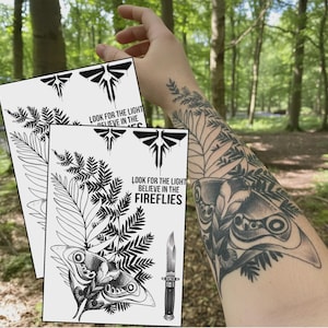 Puede incluir: Dos impresiones blancas con ilustraciones en negro de una polilla, helechos y un cuchillo. Las impresiones tienen el texto "LOOK FOR THE LIGHT BELIEVE IN THE FIREFLIES". Un tatuaje de una polilla y helechos es visible en el brazo.